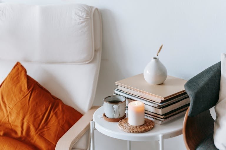 Cup of tea and flaming candle against heap of books with dry spike in vase between armchairs at home on white background