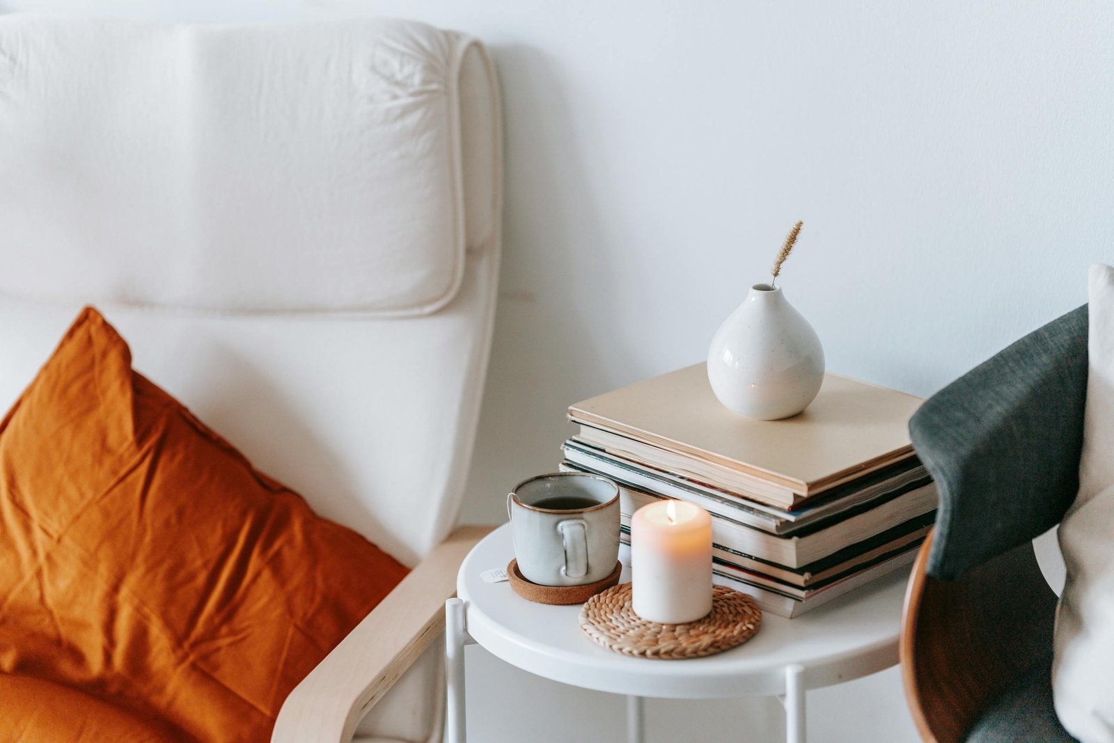 Cup of tea and flaming candle against heap of books with dry spike in vase between armchairs at home on white background