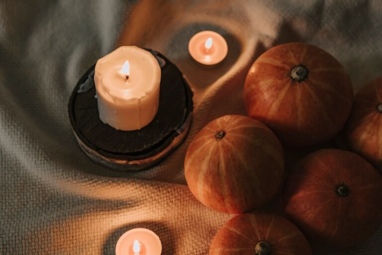 Lit autumn candle with warm amber glow on wooden table beside fall leaves and blanket