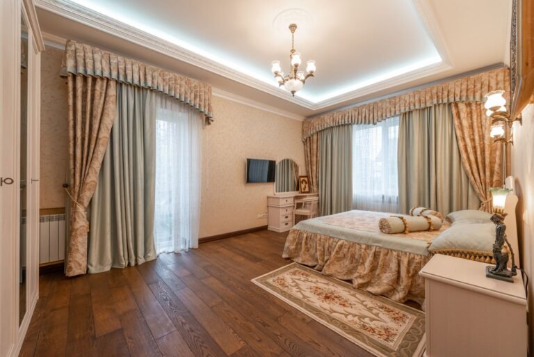 Cozy bedroom with layered area rug on hardwood floor next to white bedding and natural light